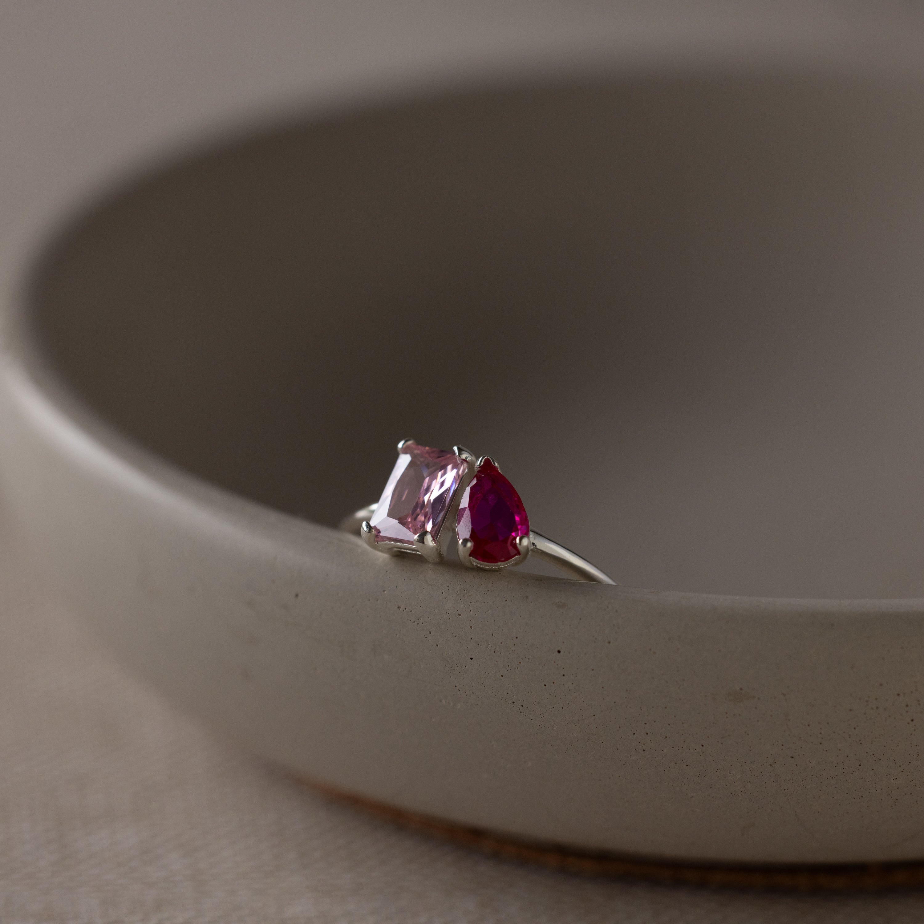 The Toi et Moi Birthstone Ring in Sterling Silver, with one pink and one red gemstone, sits on the edge of a round ceramic bowl.
