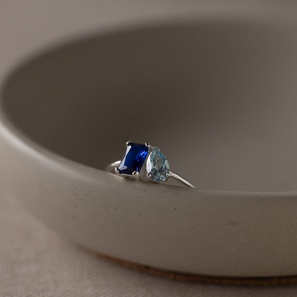 The Toi et Moi Birthstone Ring in Sterling Silver features a blue rectangular gem and a light blue teardrop, sparkling atop a beige dish—an elegant and customizable Christmas gift.