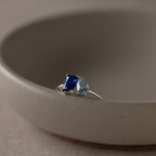 The Toi et Moi Birthstone Ring in Sterling Silver features a blue rectangular gem and a light blue teardrop, sparkling atop a beige dish—an elegant and customizable Christmas gift.