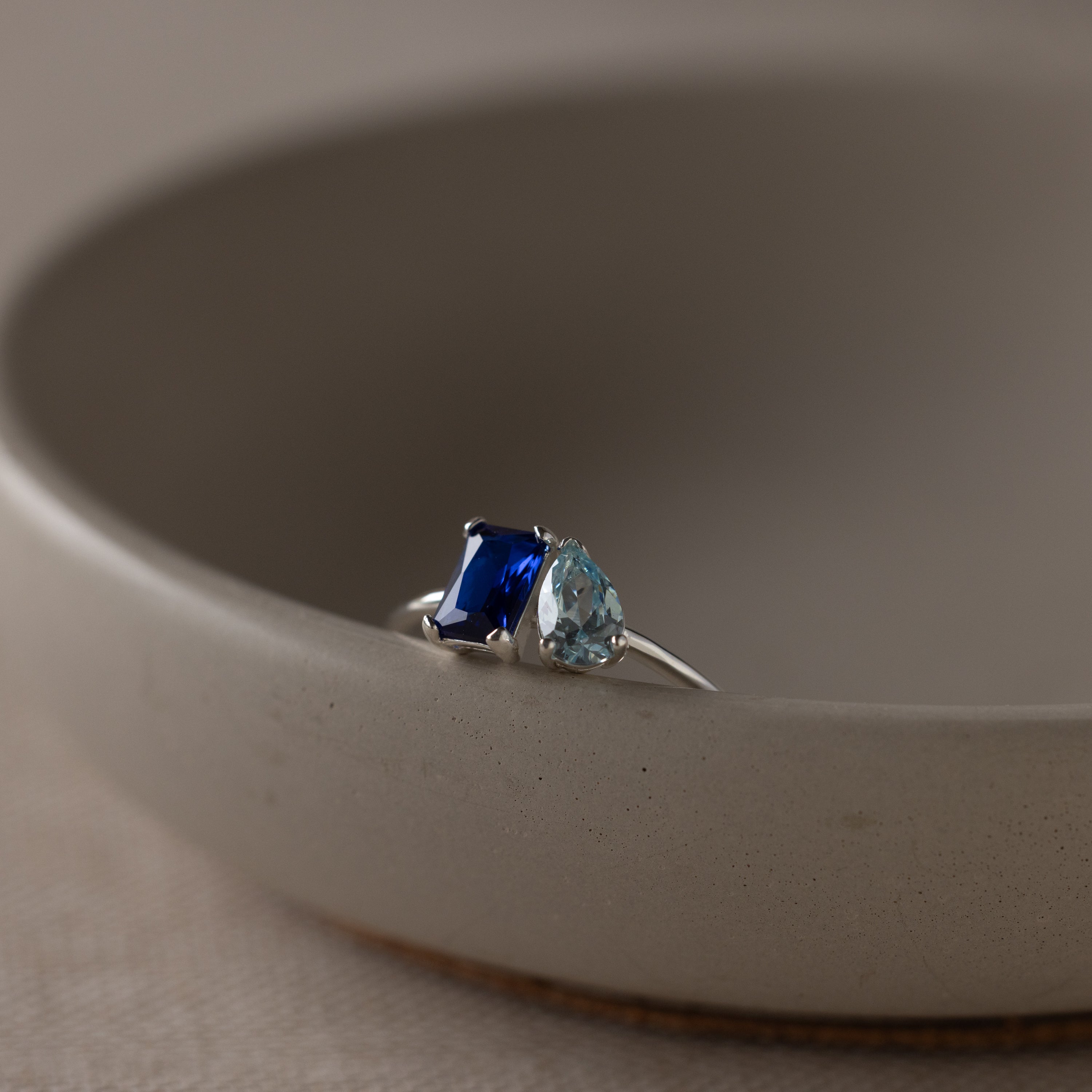 The Toi et Moi Birthstone Ring in Sterling Silver features a blue rectangular gem and a light blue teardrop, sparkling atop a beige dish—an elegant and customizable Christmas gift.