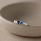 The Cluster Baguette Birthstone Ring in Sterling Silver, with pink, blue, and teal gemstones, rests on the rim of a beige ceramic bowl.
