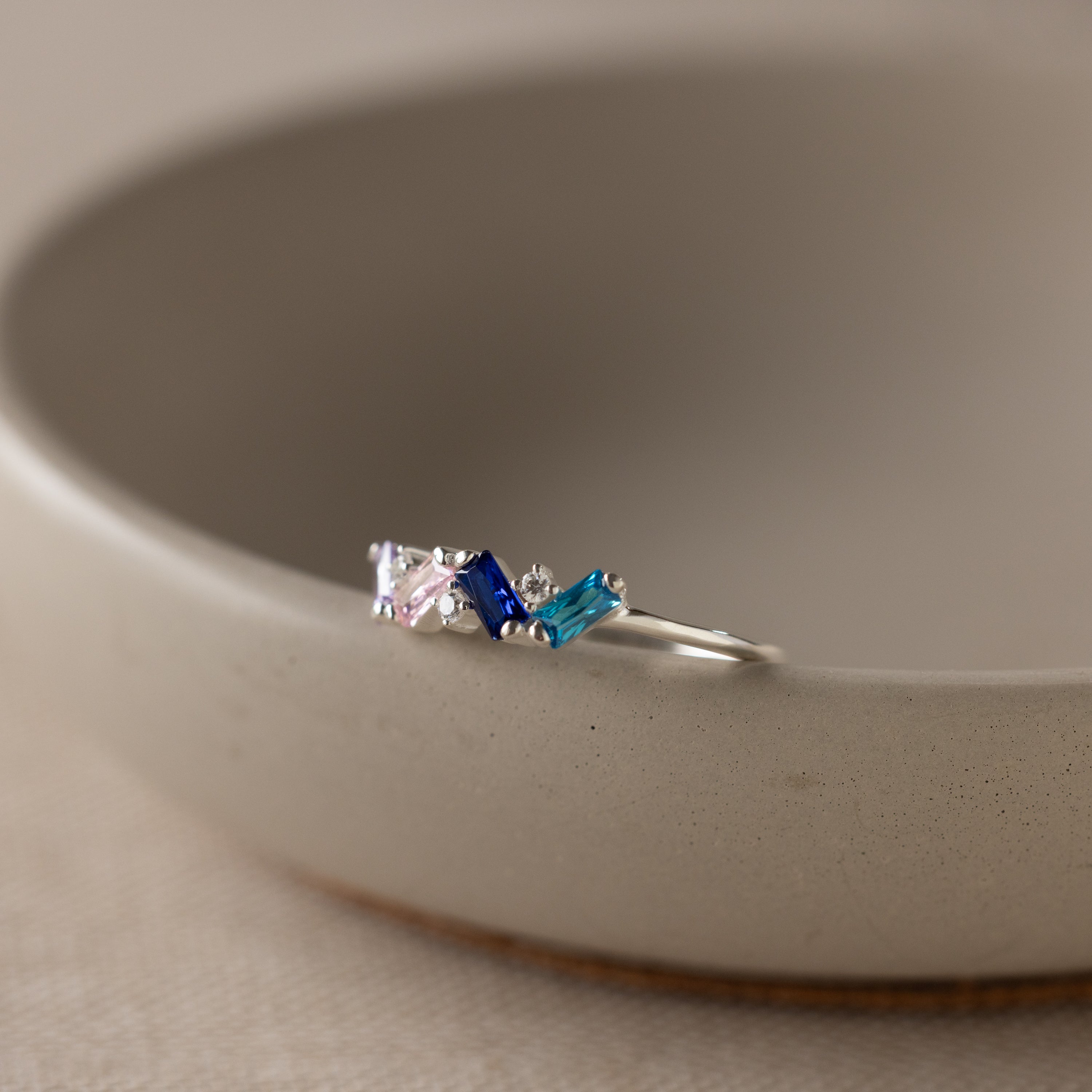 The Cluster Baguette Birthstone Ring in Sterling Silver, with pink, blue, and teal gemstones, rests on the rim of a beige ceramic bowl.