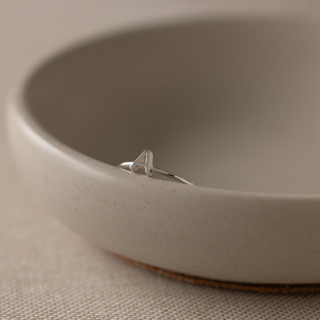 The Alex Dainty Letter Ring in Sterling Silver, featuring an A initial, sits on the rim of a beige ceramic bowl, showcasing the elegant charm of personalized jewelry.