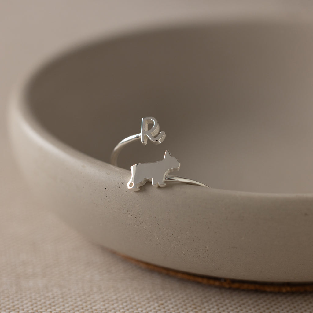 The Duo Pet Initial Wrap Ring, featuring a silver dog shape and the letter R, rests on the edge of a beige ceramic bowl.