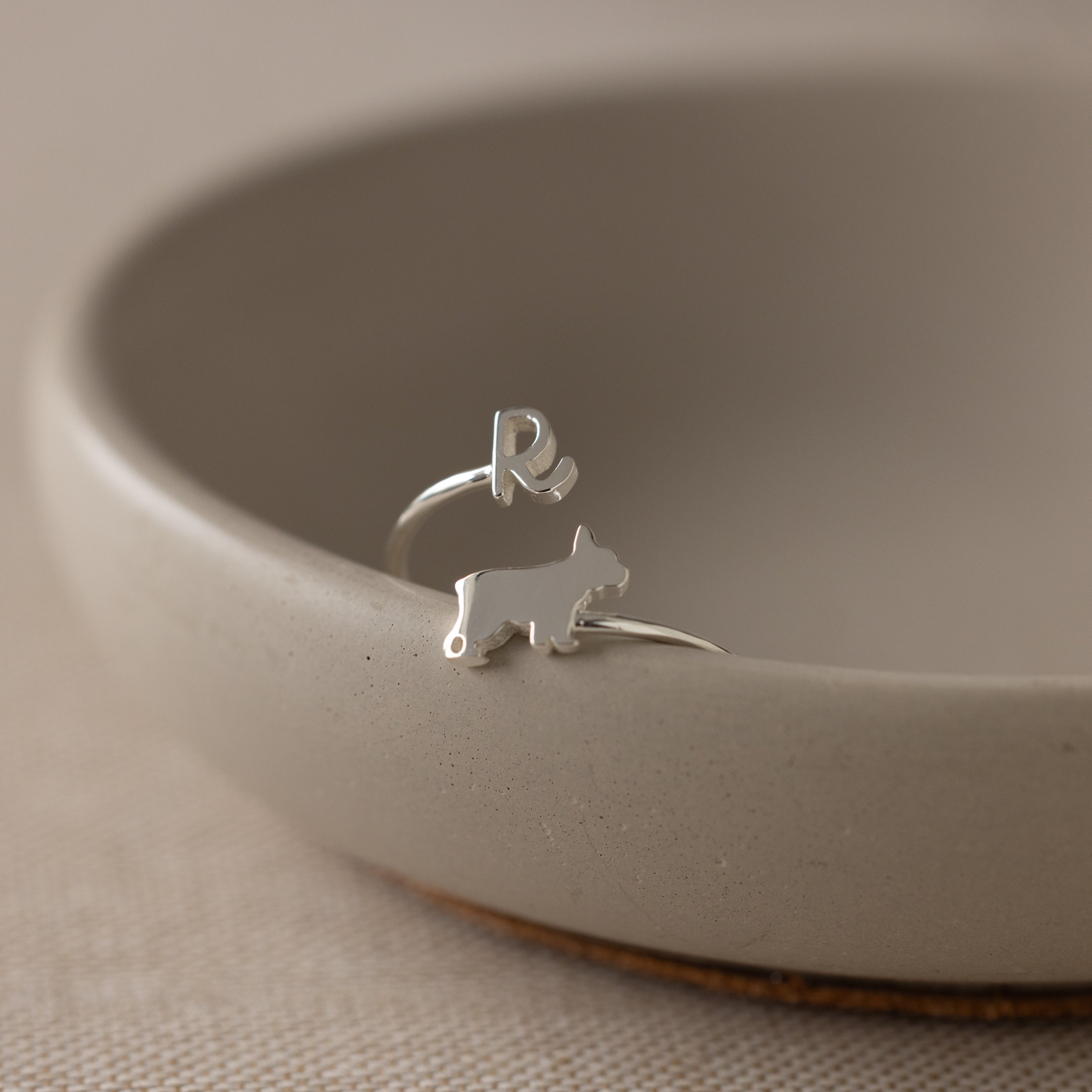 The Duo Pet Initial Wrap Ring, featuring a silver dog shape and the letter R, rests on the edge of a beige ceramic bowl.