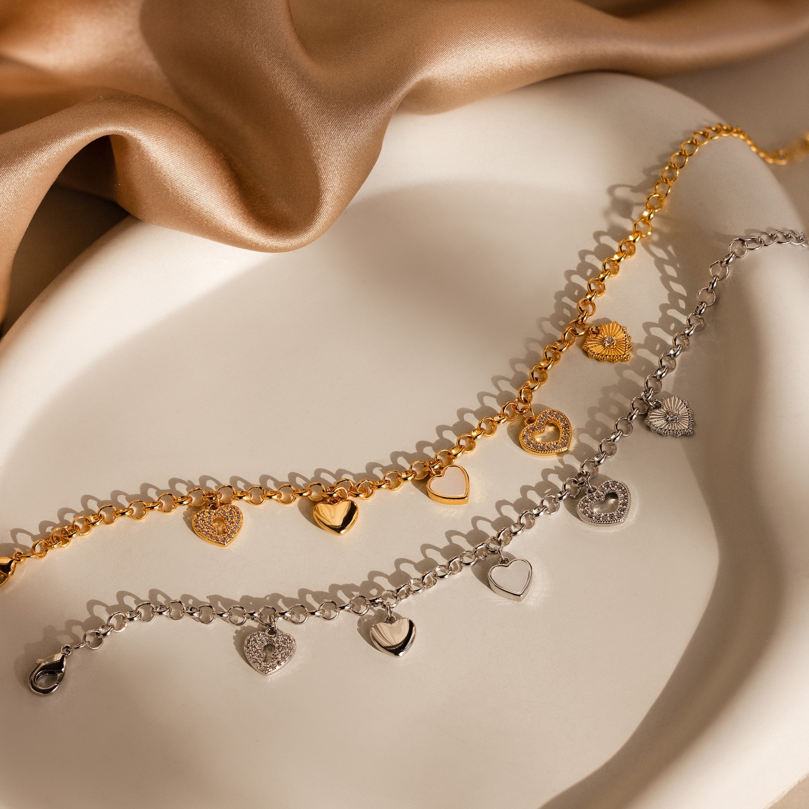 The Heart Charm Bracelet features gold and silver heart pendants, displayed elegantly on a white dish with a satin cloth background—an ideal gift for your girlfriend.
