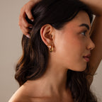 Woman with long dark hair wearing Spiral Swirl Hoops in 18K Gold, tilting her head with a hand behind her ear against a neutral background—perfect boho vacation jewelry.