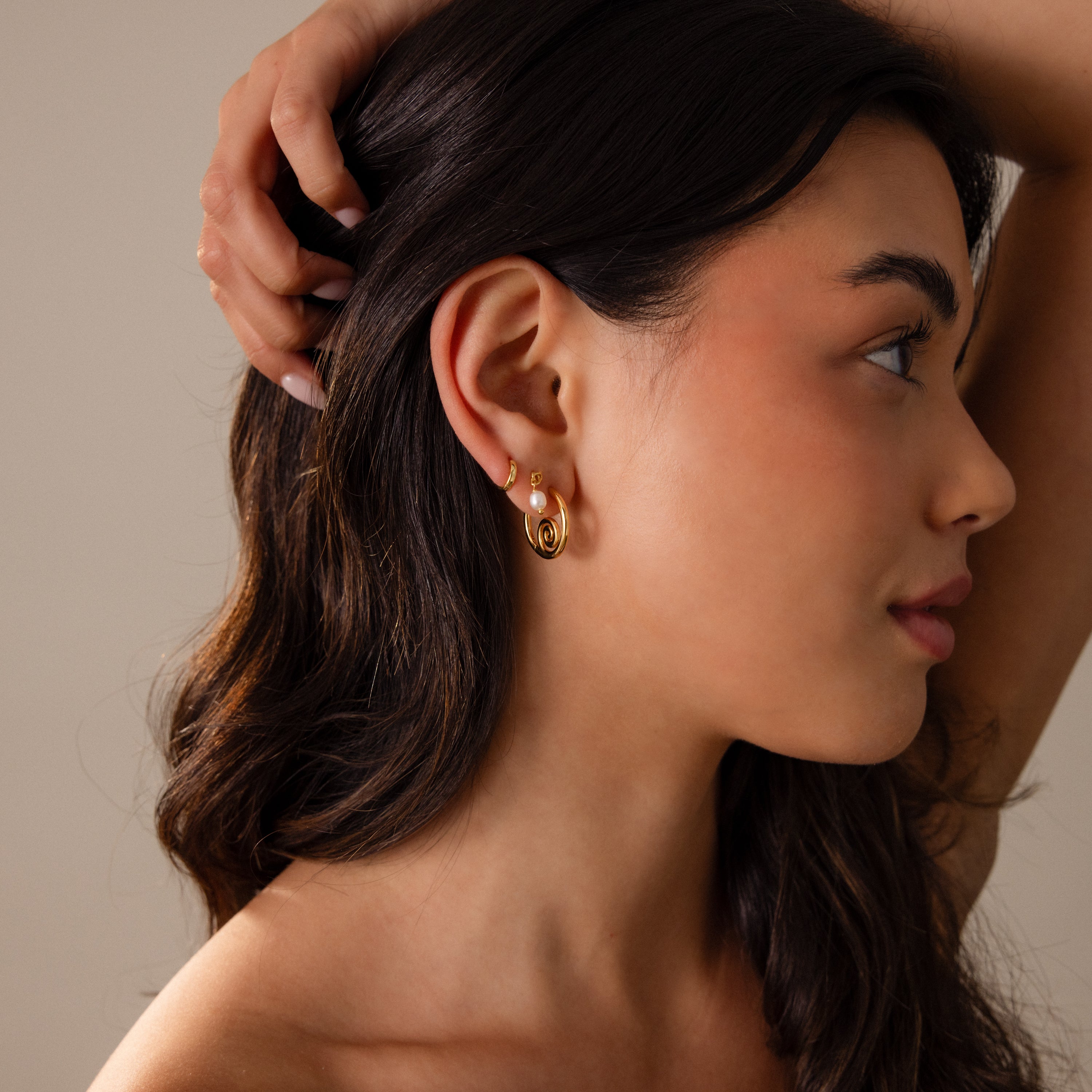 Woman with long dark hair wearing Spiral Swirl Hoops in 18K Gold, tilting her head with a hand behind her ear against a neutral background—perfect boho vacation jewelry.