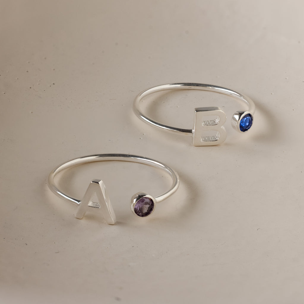 Two Alex Initial Birthstone Rings in sterling silver—one with ‘A’ and a purple gem, the other with ‘B’ and a blue gem. The perfect personalized set for a unique touch.