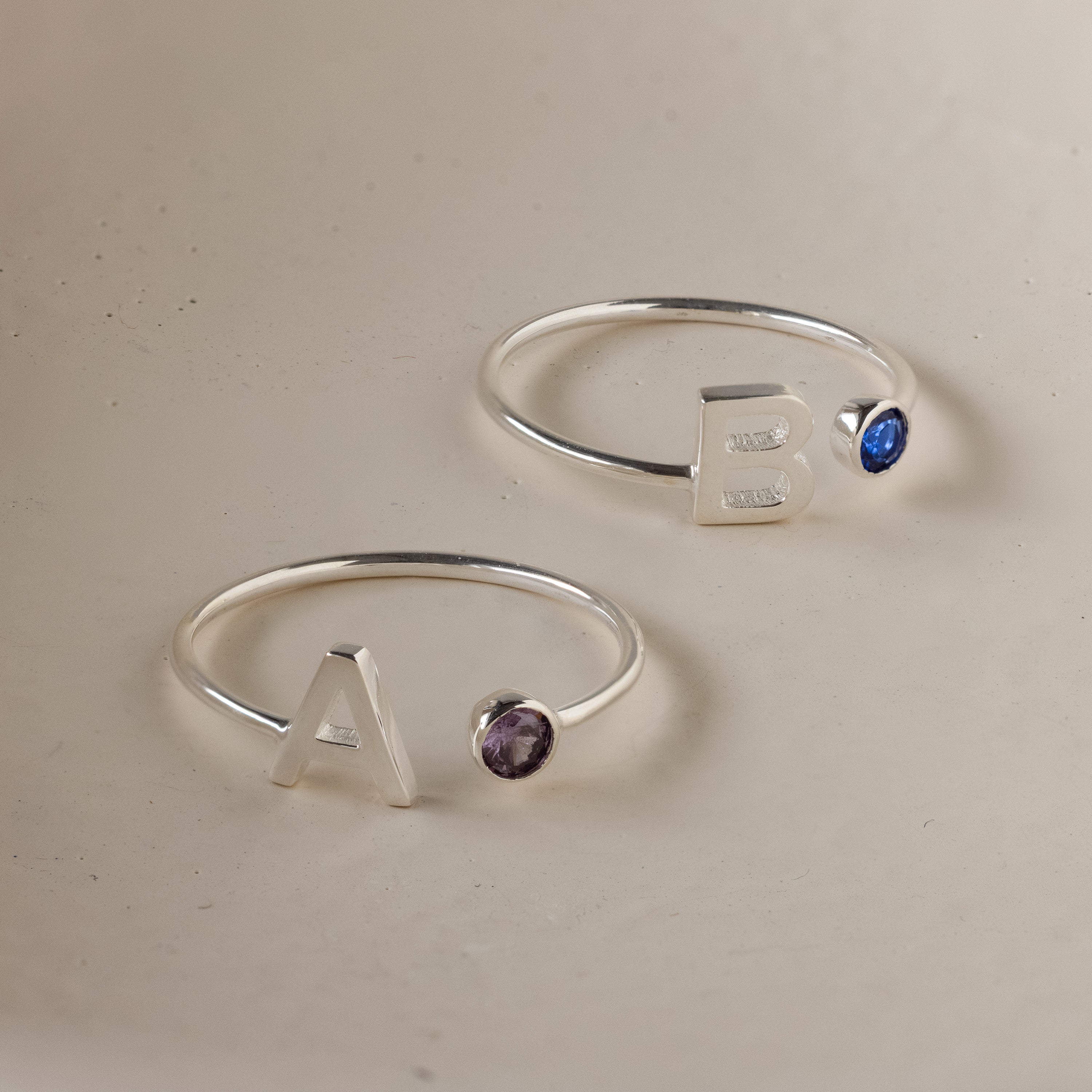 Two Alex Initial Birthstone Rings in sterling silver—one with ‘A’ and a purple gem, the other with ‘B’ and a blue gem. The perfect personalized set for a unique touch.