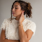 A woman with curly hair wears a textured white top and a chic accessory— the Classic Bangle in 18K Gold— as she touches her ear and appears relaxed.