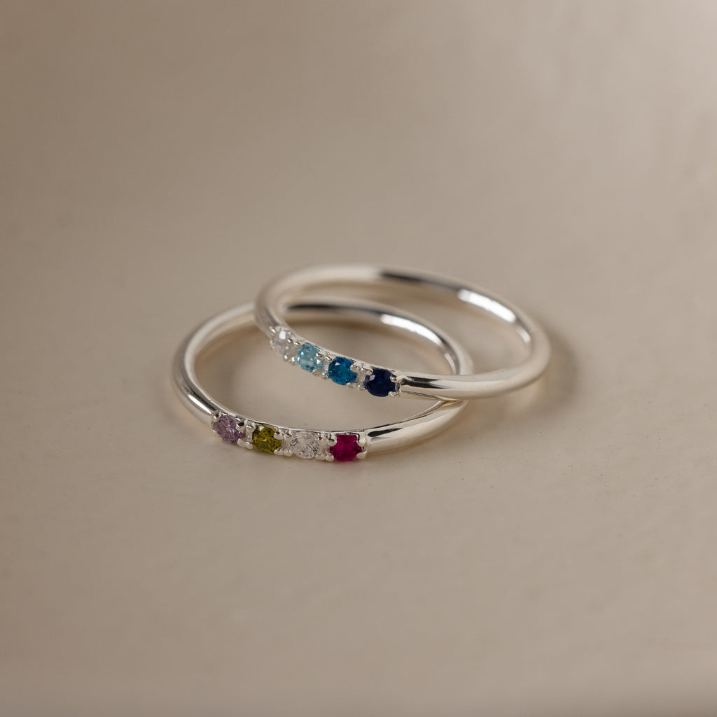 The Mini Birthstone Ring in Sterling Silver features colorful gemstones set in a row—ideal as a dainty accessory or personalized birthstone ring for any occasion, displayed on a smooth beige surface.