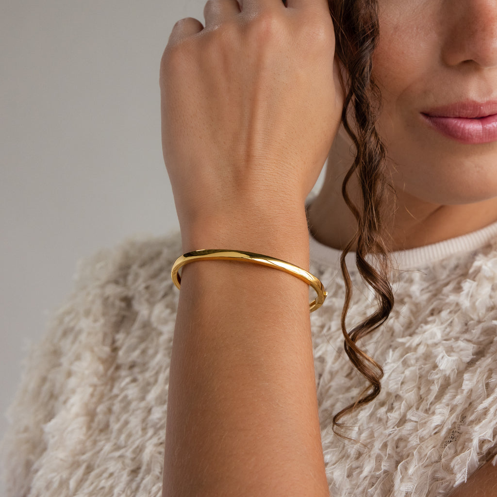 A woman showcases the Classic Bangle—a gold minimalist bracelet—her hand near her face, curly hair framing her features as she wears a textured cream top.