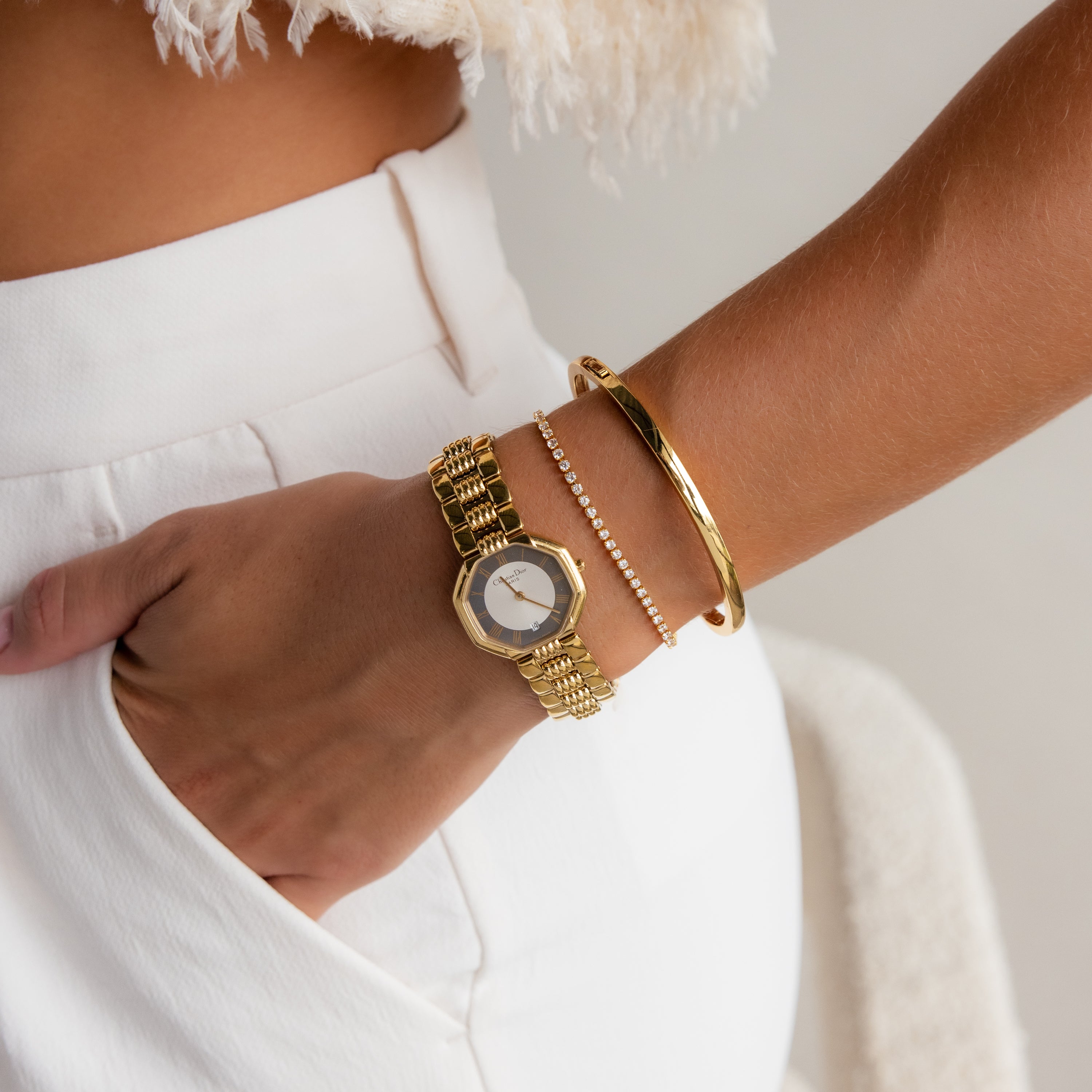 A woman in white pants wears a gold watch, the Classic Bangle in 18K Gold, and a delicate bracelet on her wrist.