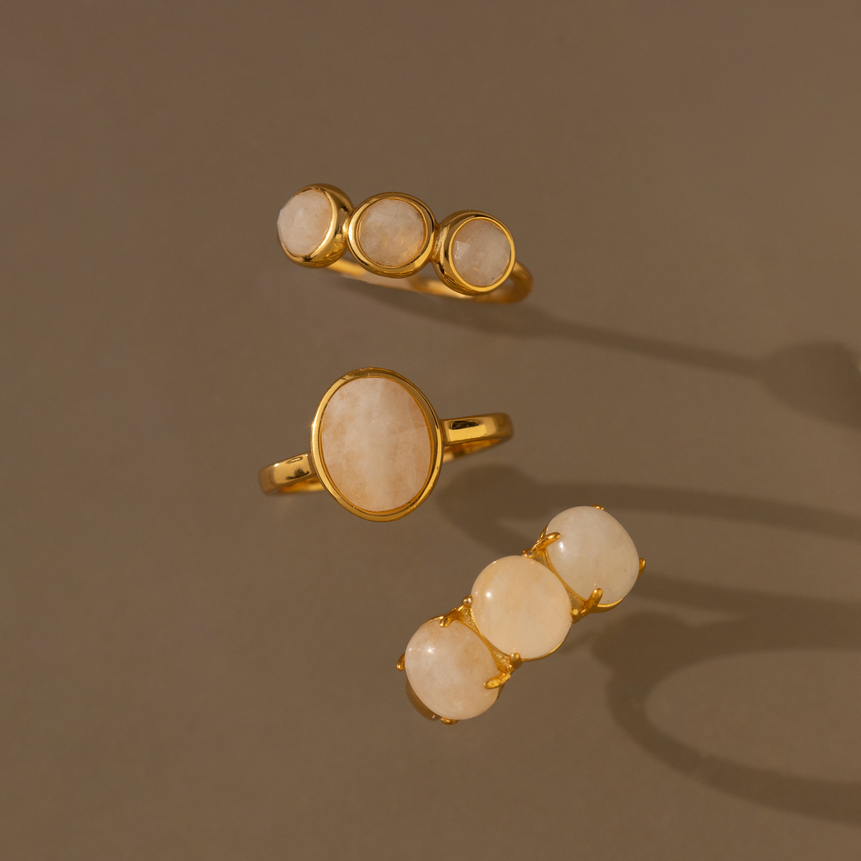 Three gold Moonstone Cluster Rings, each with oval and round light beige stones, are displayed on a brown background—an ideal choice for fans of modern moonstone jewelry.