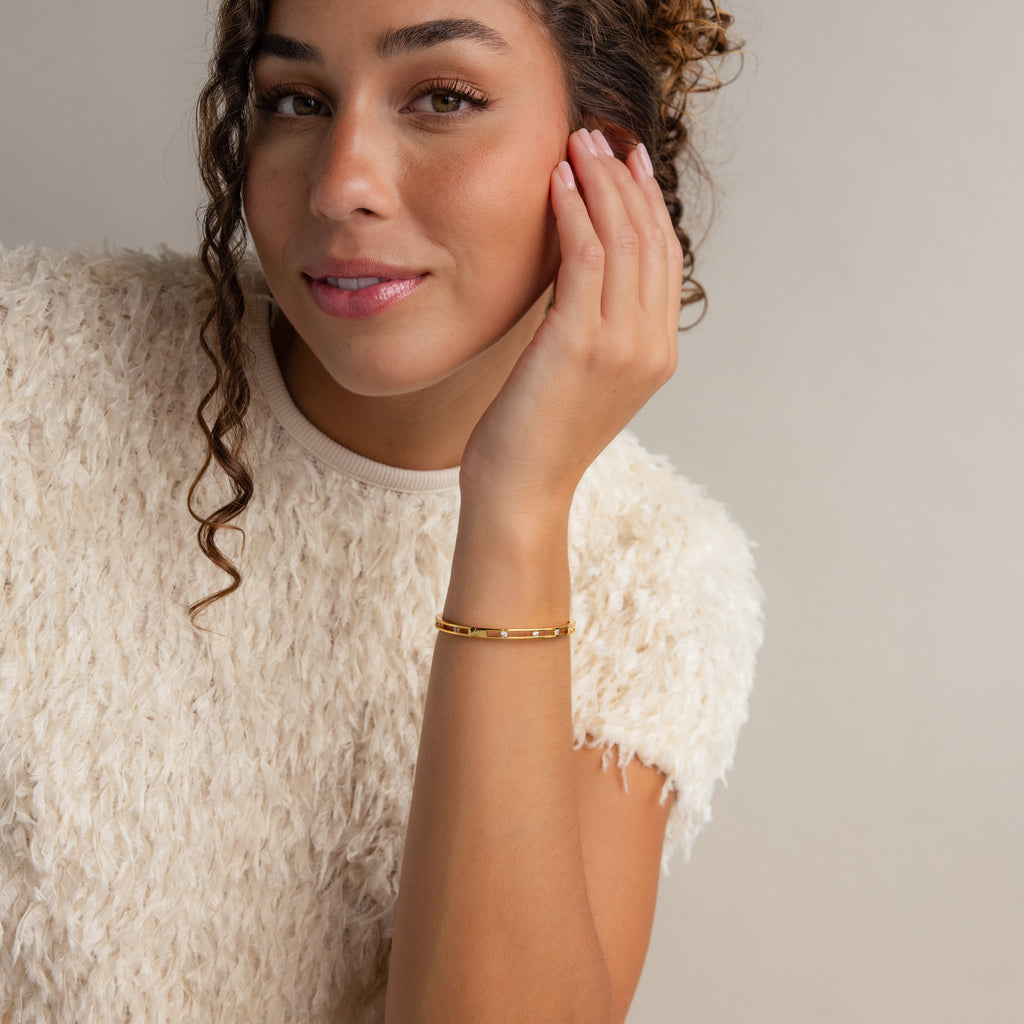 A woman with curly hair, wearing a white textured top and a sparkling Lace Diamond Bangle in 18K Gold, smiles and touches her face.