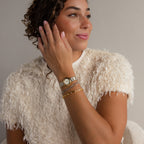 A woman with curly hair, wearing a textured white top, gold watch, and the Lace Diamond Bangle, smiles as she looks to the side.