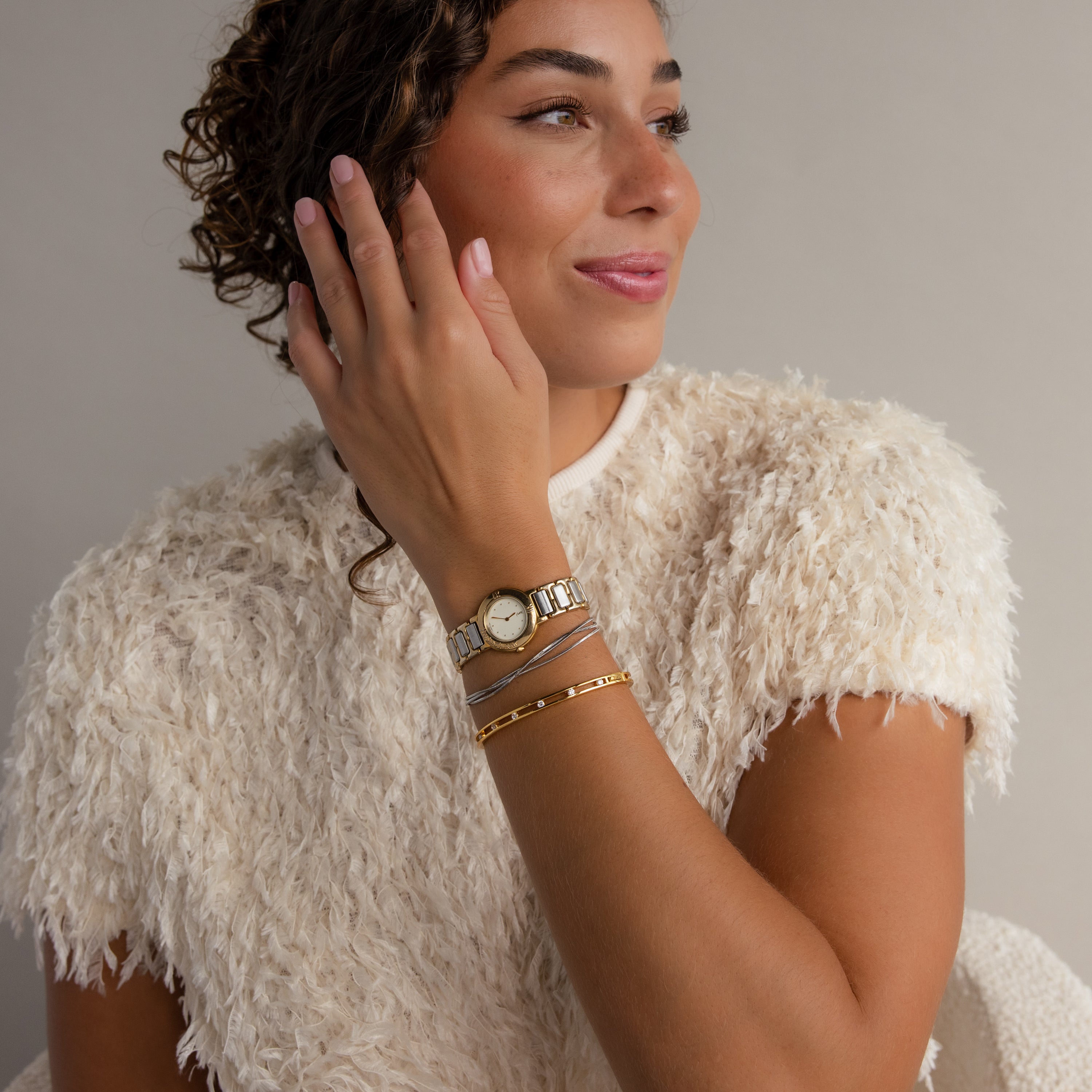 A woman with curly hair, wearing a textured white top, gold watch, and the Lace Diamond Bangle, smiles as she looks to the side.