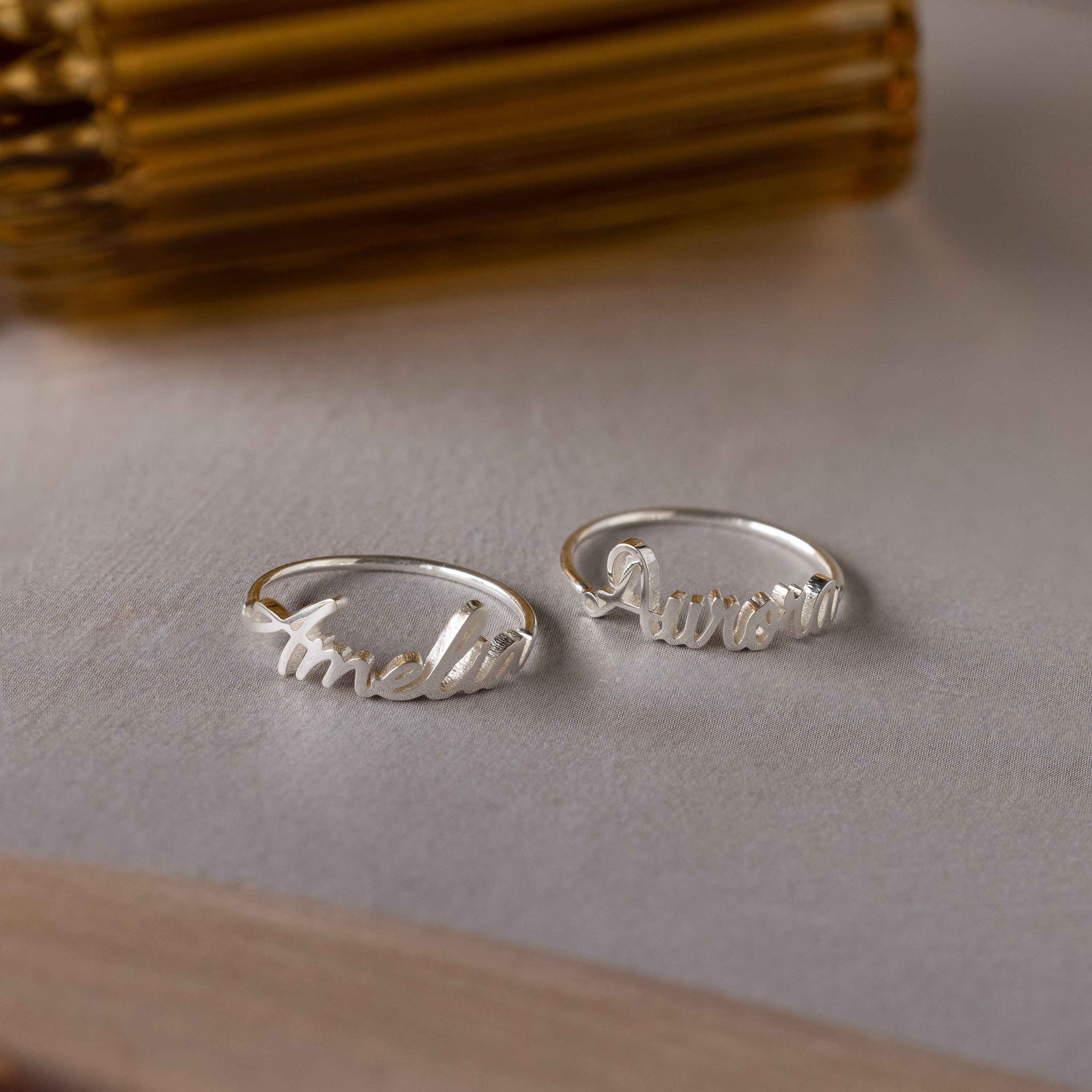 Two Grace Script Name Rings in Sterling Silver engraved with Amelia and Aurora in elegant script, displayed on a light surface—an ideal personalized ring for any occasion.