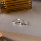 Two sterling silver rings rest on the table—one engraved with "Oliver," the other is the Flower Bar Ring, a skinny band featuring a botanical jewelry leaf design.