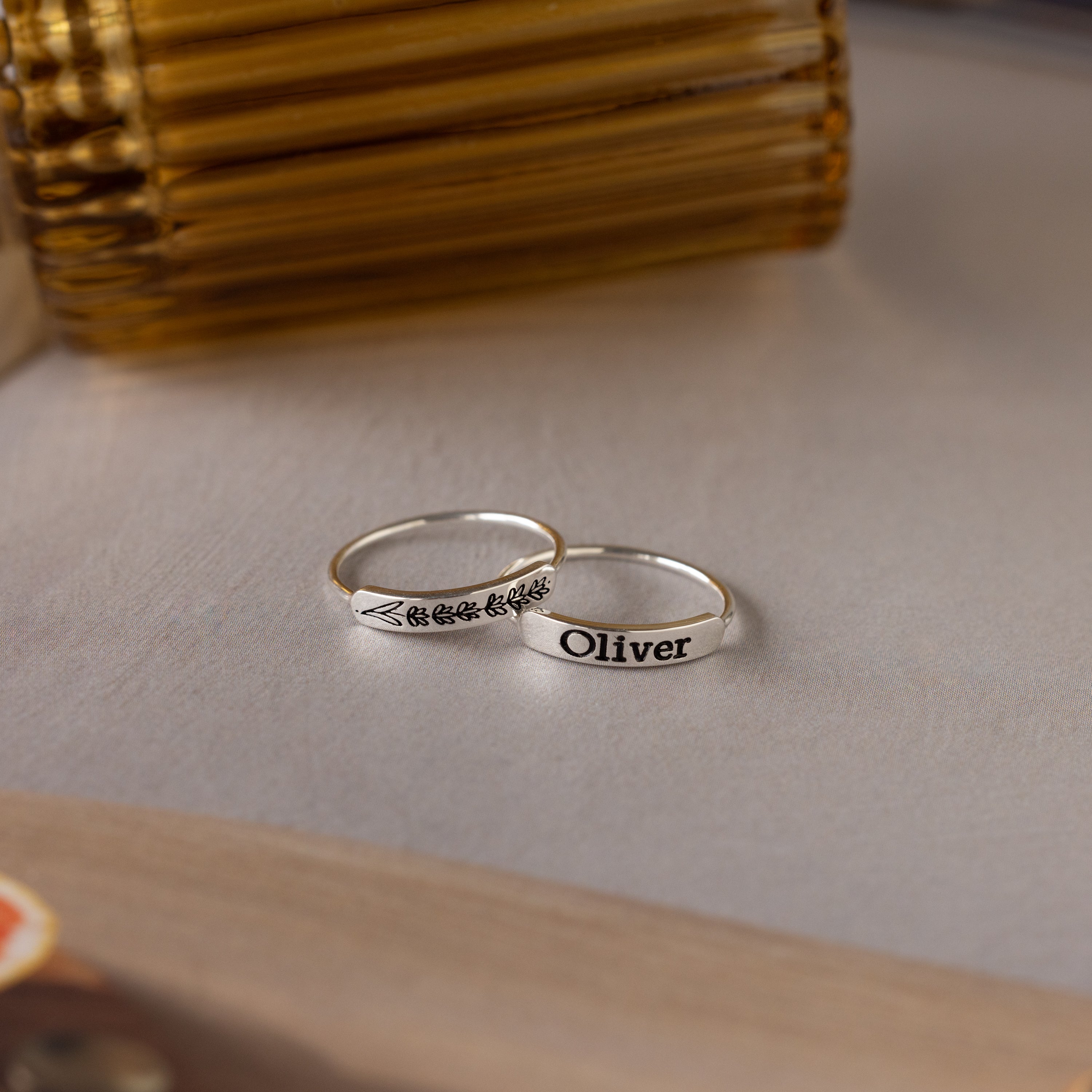 Two sterling silver rings rest on the table—one engraved with "Oliver," the other is the Flower Bar Ring, a skinny band featuring a botanical jewelry leaf design.