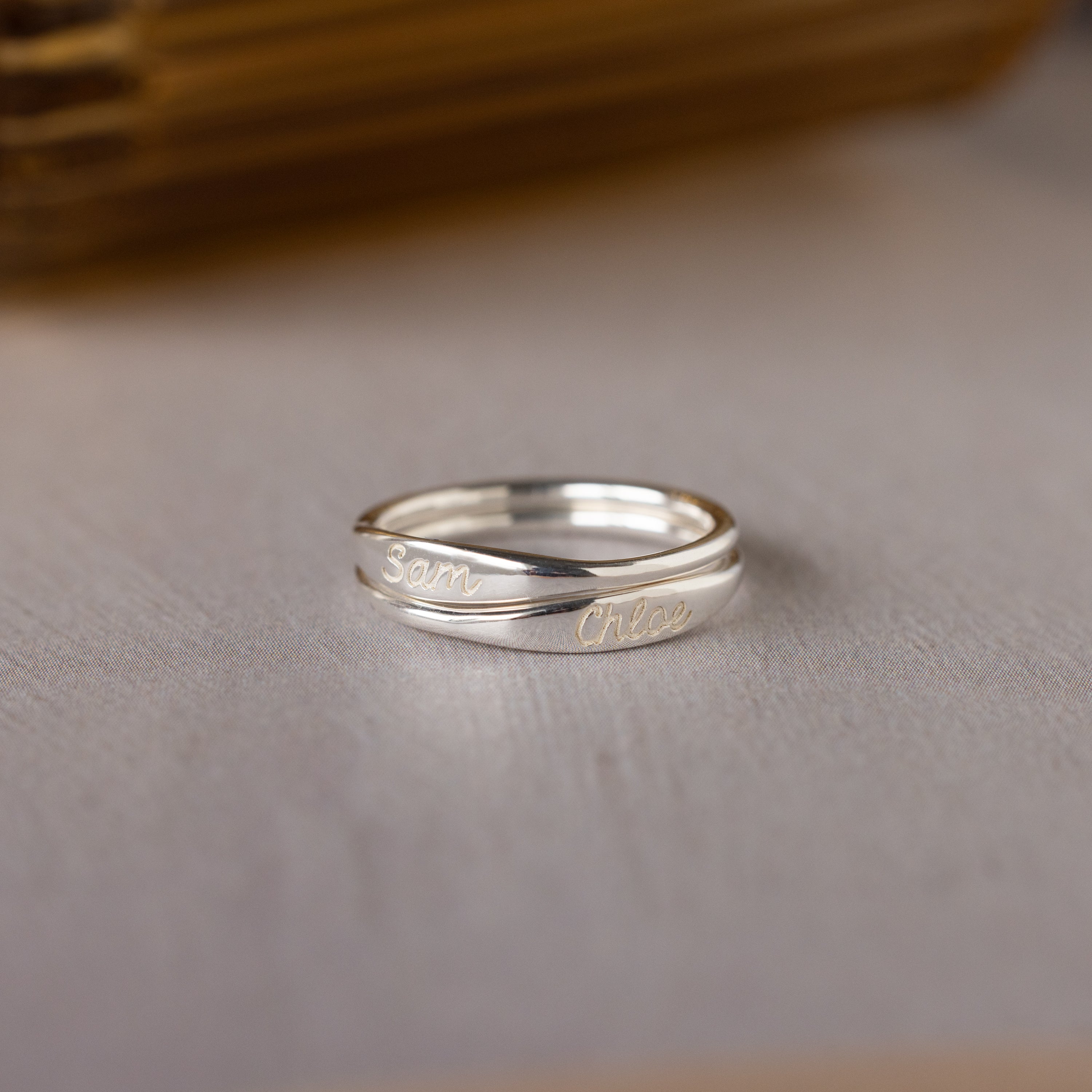 The Ann Skinny Name Ring in Sterling Silver, engraved with names like Sam and Chloe, rests on a light surface—ideal for stacking or as a thoughtful Christmas gift for her.