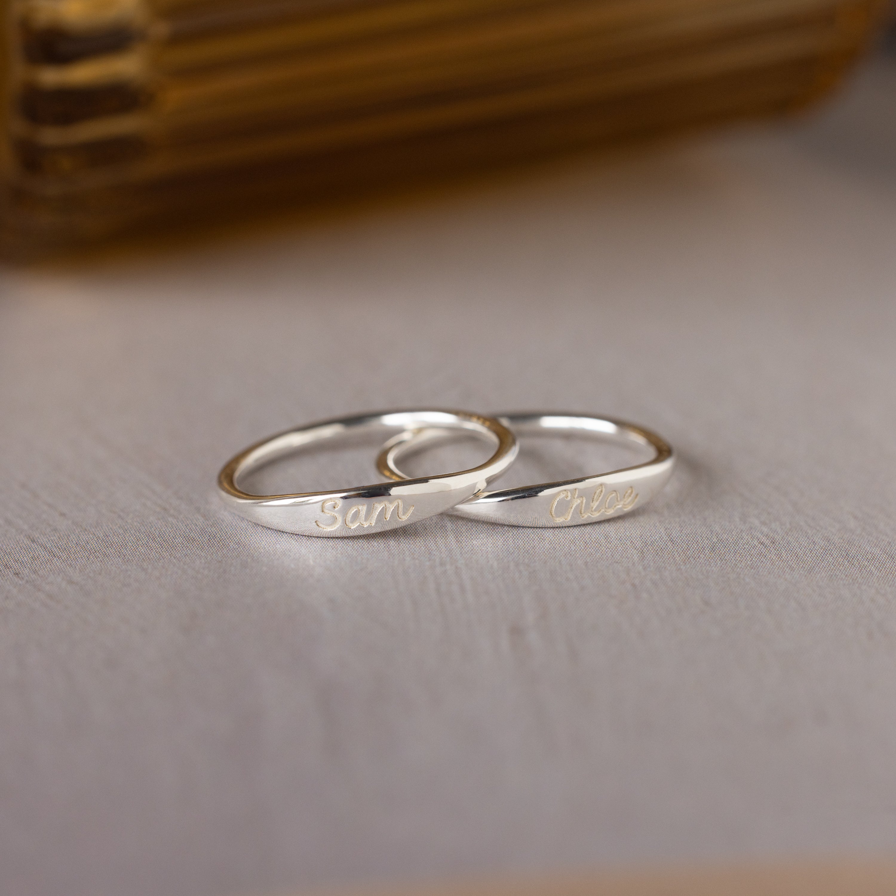Two Ann Skinny Name Rings in Sterling Silver, engraved with "Sam" and "Chloe", rest on a light wooden surface—a thoughtful Christmas gift for her.