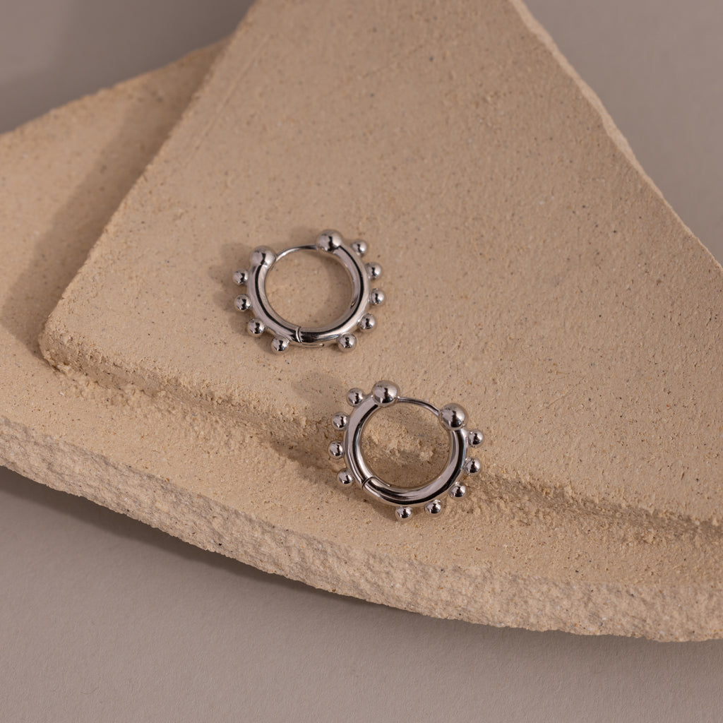 Beaded Hoops in Sterling Silver, adorned with small round accents and displayed on textured beige stone slabs, capture a subtle, textured glow.