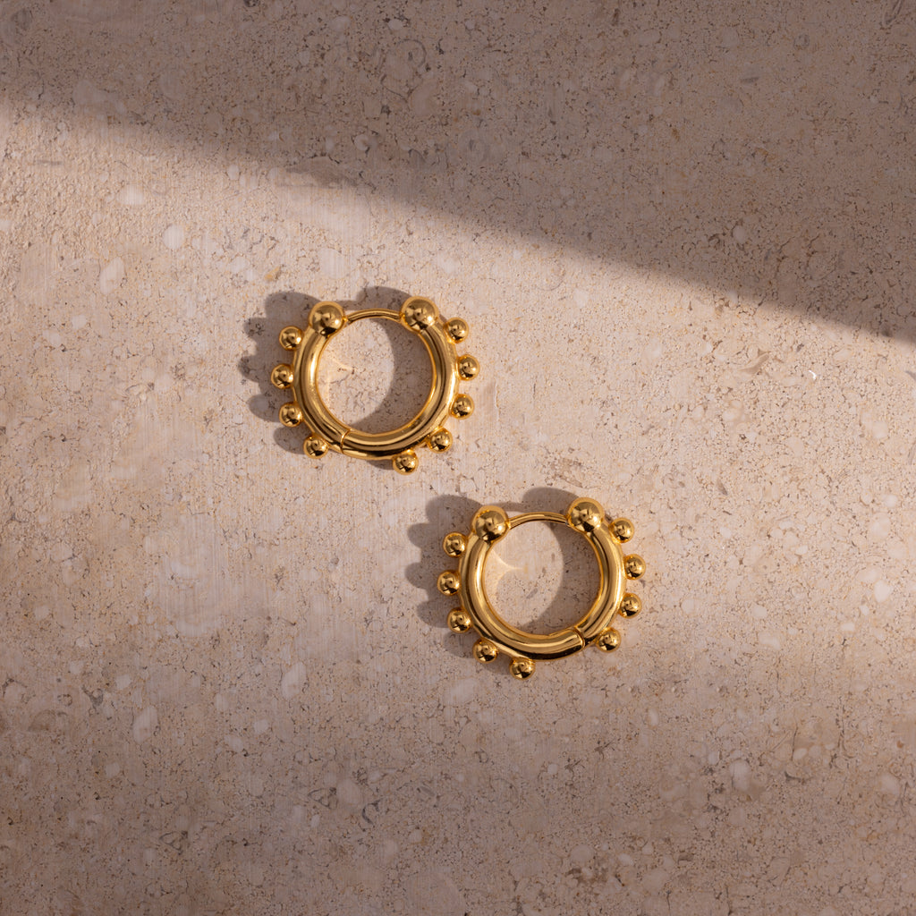 A pair of Beaded Hoops in 18K Gold with polished beads, displayed on a textured stone surface with interplay of light and shadow.