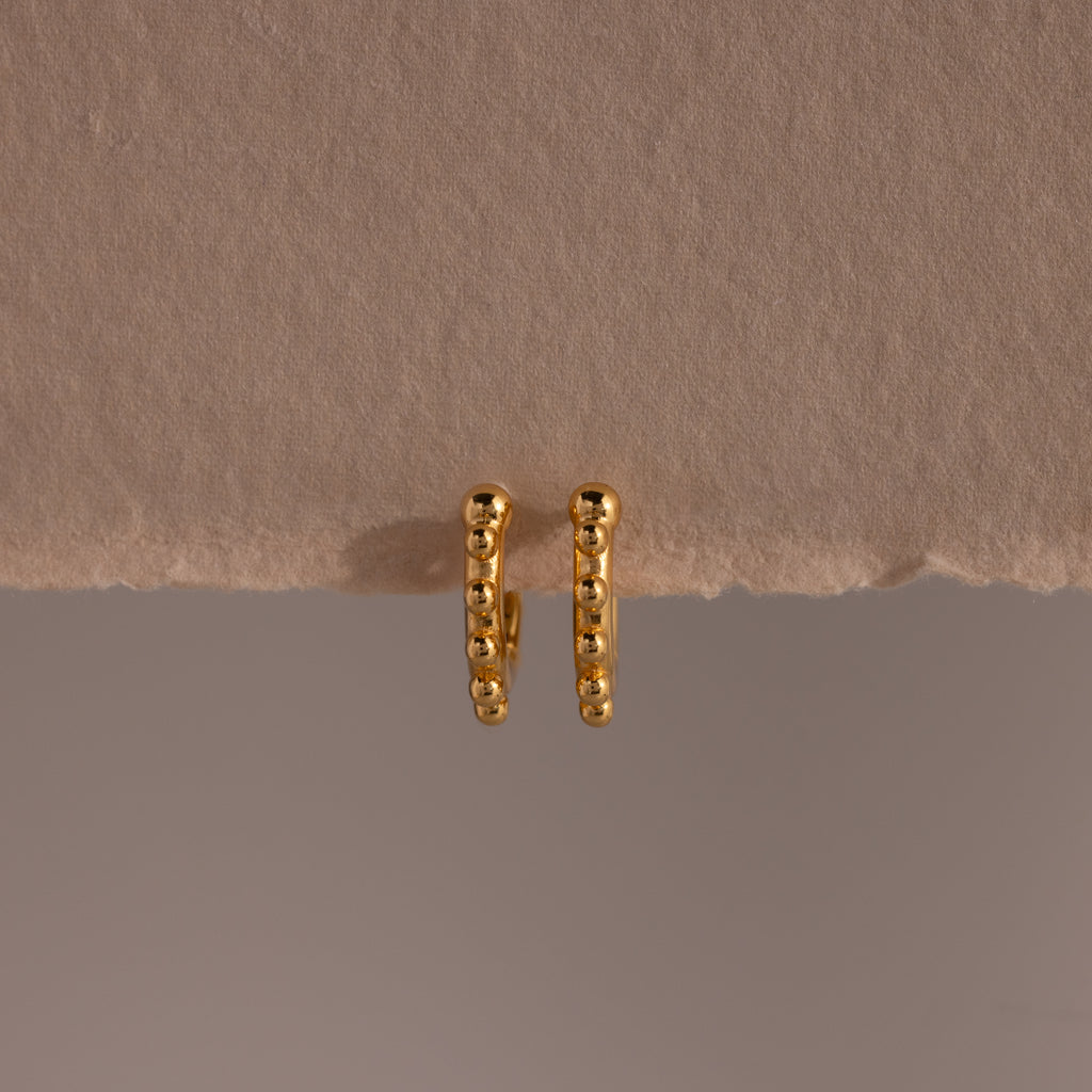 A pair of Beaded Hoops in 18K Gold, showcasing polished beads and a beaded texture, displayed against a beige textured surface.