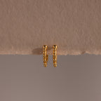A pair of Beaded Hoops in 18K Gold, showcasing polished beads and a beaded texture, displayed against a beige textured surface.
