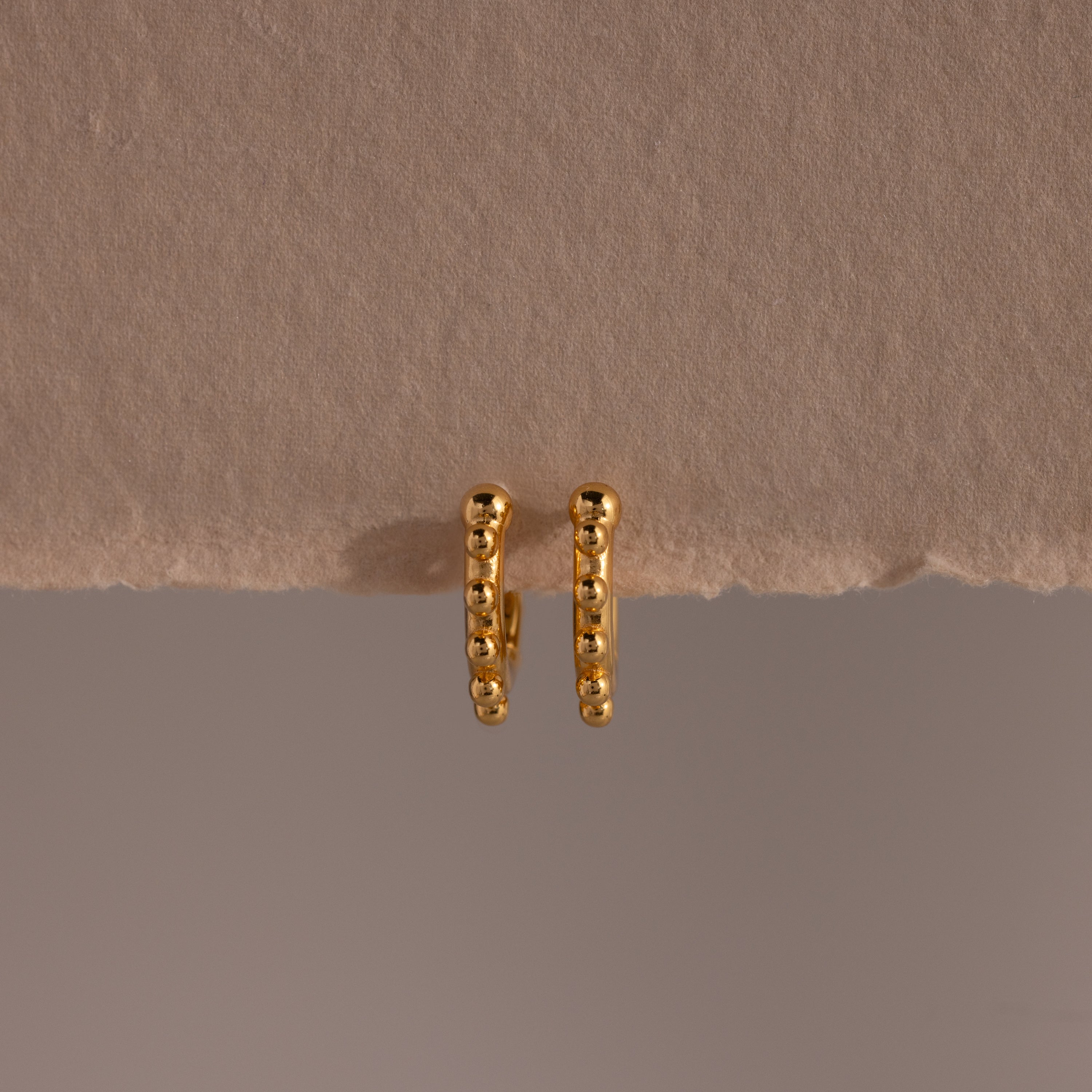 A pair of Beaded Hoops in 18K Gold, showcasing polished beads and a beaded texture, displayed against a beige textured surface.