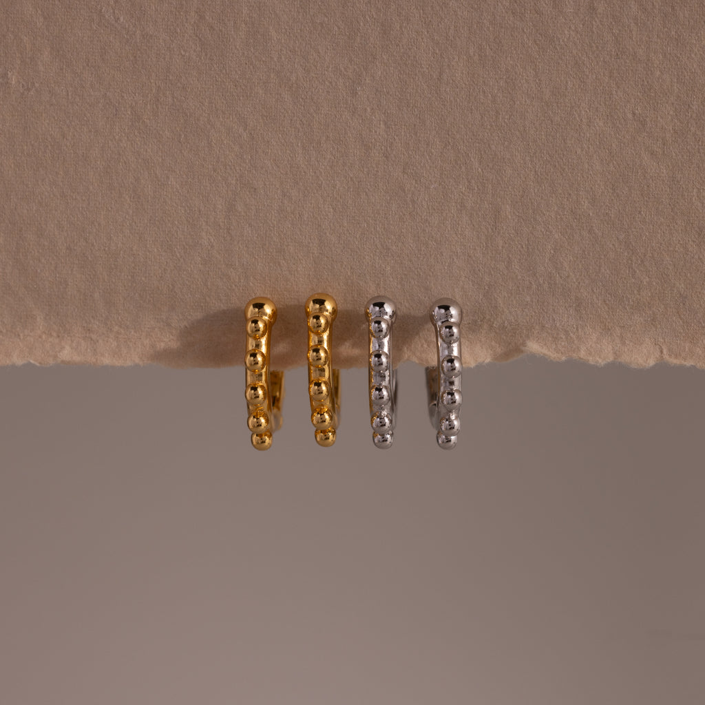 Two pairs of Beaded Hoops—one gold, one silver—are displayed on a beige textured surface. Each pair features polished beads for a refined, modern twist on classic hoop earrings.