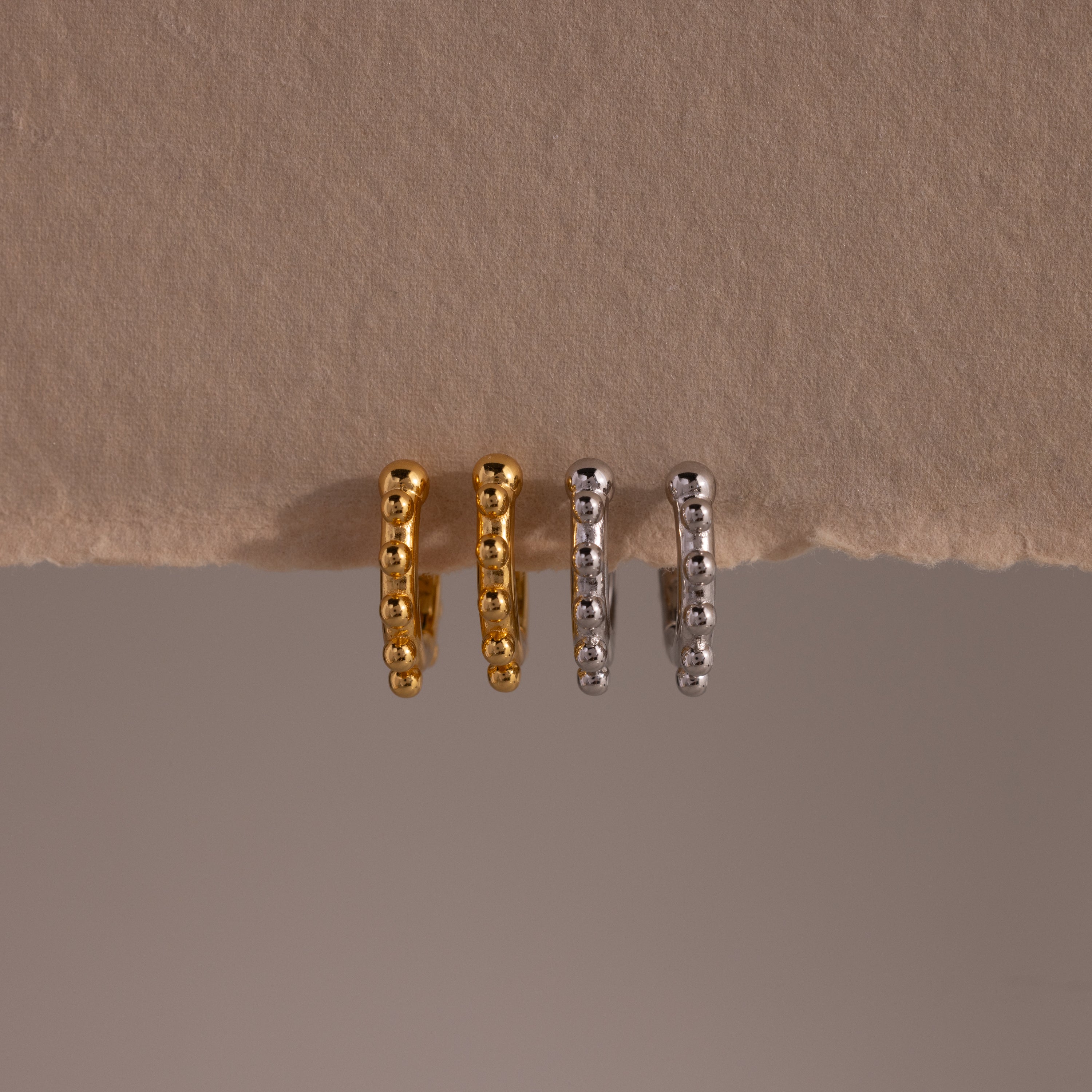 Two pairs of Beaded Hoops—one gold, one silver—are displayed on a beige textured surface. Each pair features polished beads for a refined, modern twist on classic hoop earrings.