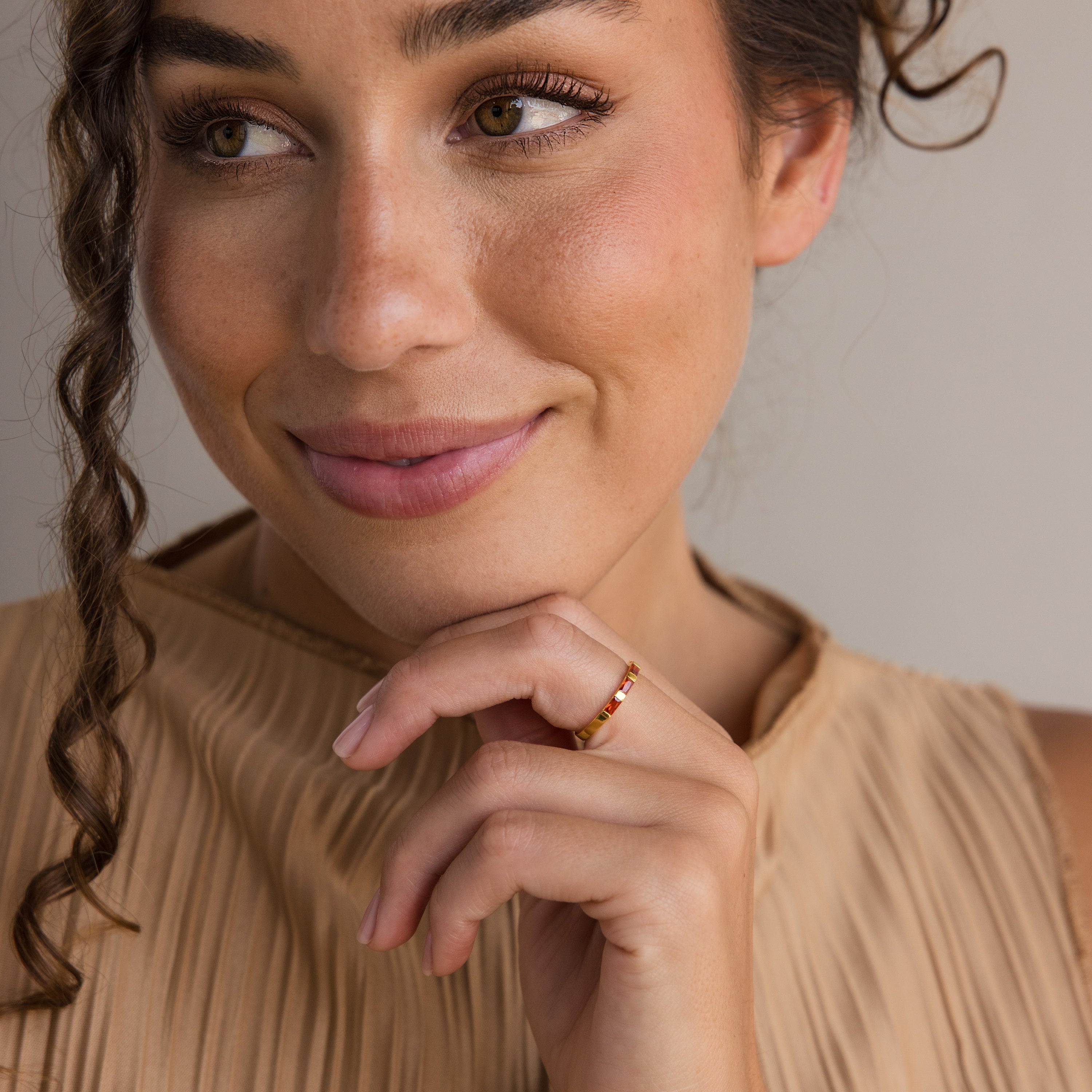 A smiling woman in a pleated beige top touches her chin, wearing the Half Eternity Birthstone Ring—a gold band featuring a striking red baguette-cut gemstone.