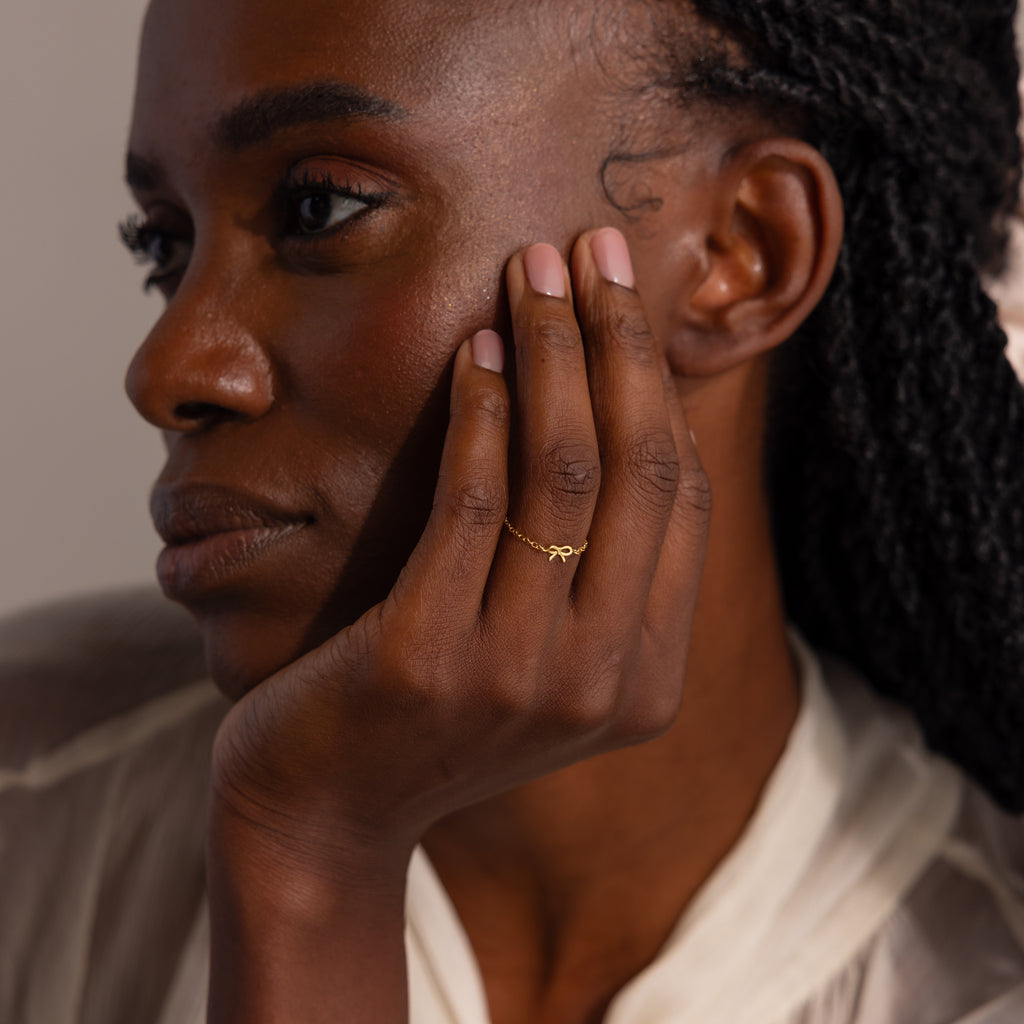 Close-up portrait of model showcasing a delicate gold chain ring with a small bow charm on her finger.