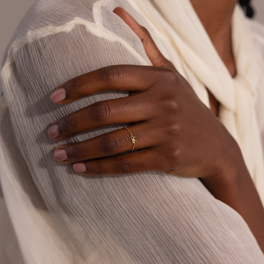 Close-up of model wearing a delicate gold chain ring with a small bow charm, styled against a sheer, ivory blouse.