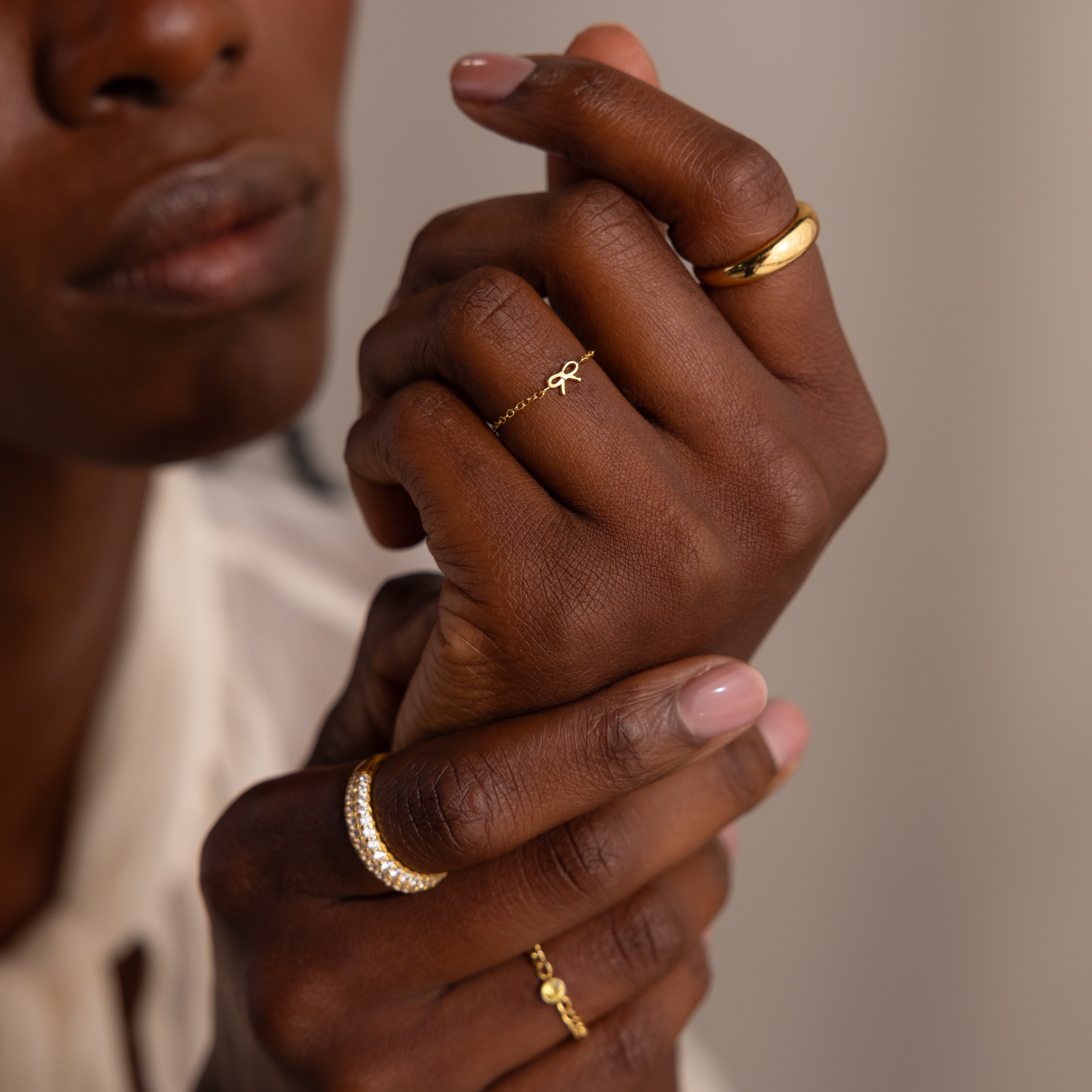 A close-up of model's hands showcasing a collection of gold rings, with the focal point being a delicate chain ring featuring a tiny gold bow.
