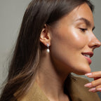 A woman with long brown hair smiles and touches her chin while wearing a tan blazer and Pearl Diamond Drop Earrings.