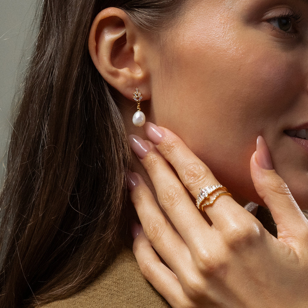 A woman wearing Pearl Diamond Drop Earrings touches her face, showcasing her manicured nails and gold rings.