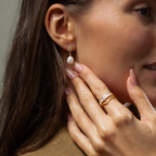A woman wearing Pearl Diamond Drop Earrings touches her face, showcasing her manicured nails and gold rings.