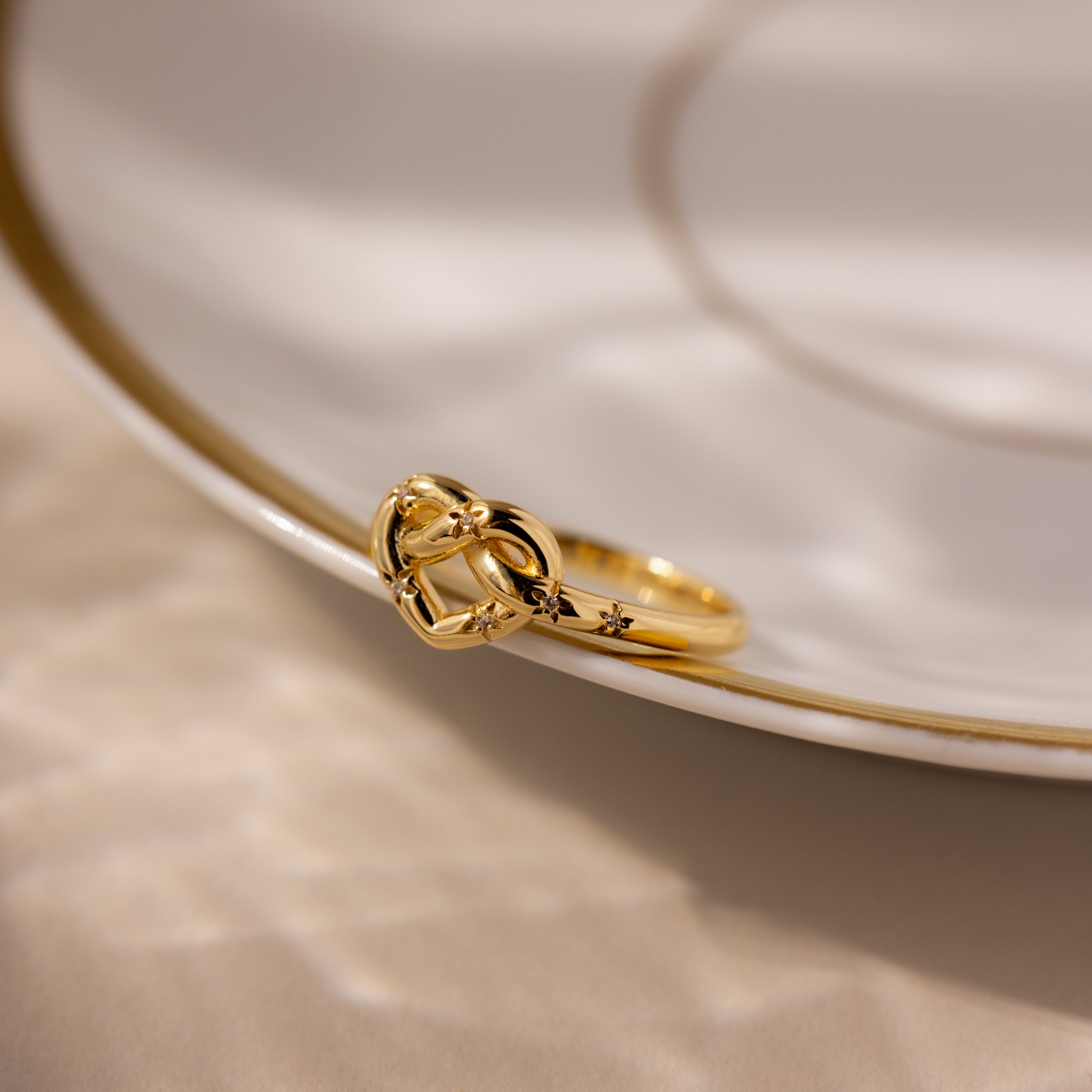 The Starry Love Knot Ring sits elegantly on the edge of a white plate adorned with a gold rim.