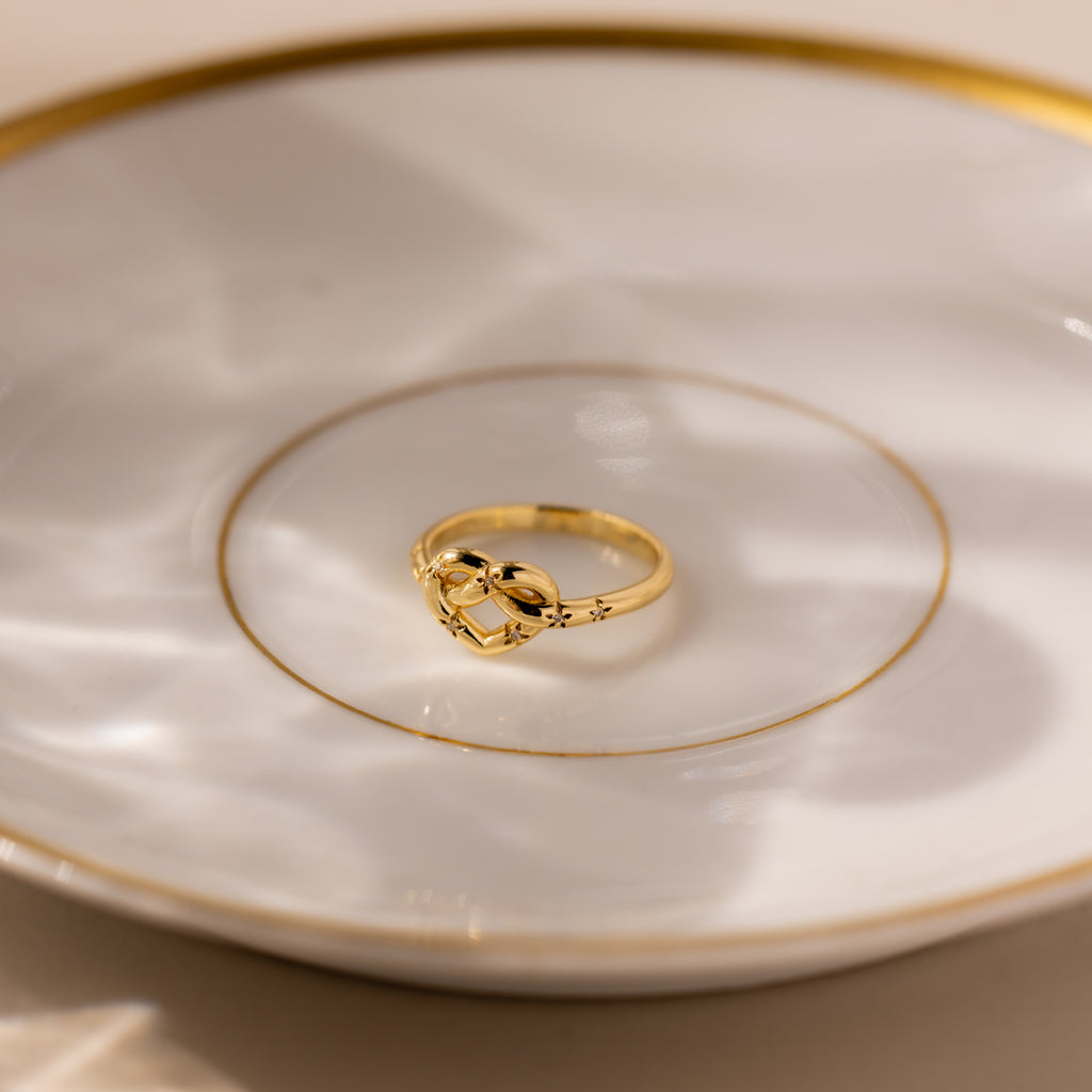 The Starry Love Knot Ring, featuring a heart-shaped design, rests on a white plate adorned with gold trim and circular patterns.