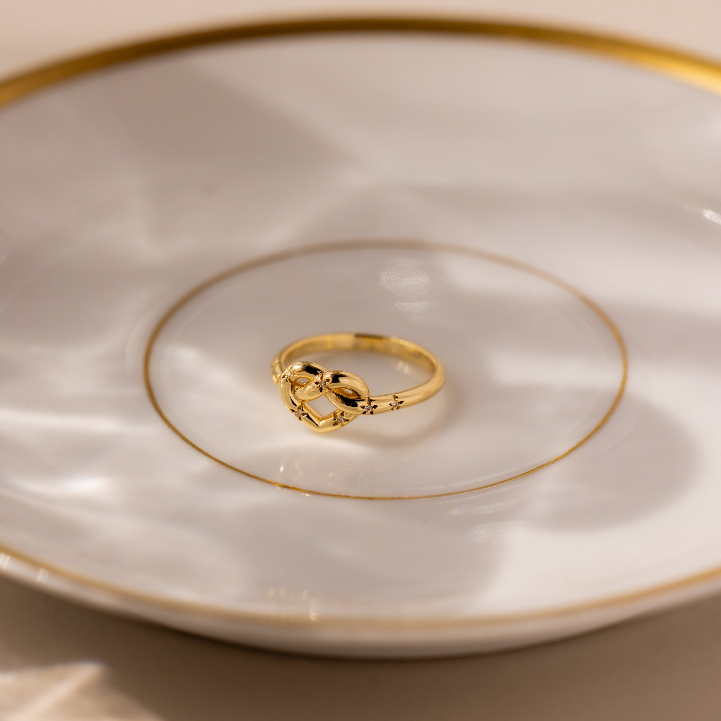 The Starry Love Knot Ring, featuring a heart-shaped design, rests on a white plate adorned with gold trim and circular patterns.