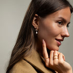 Woman in a tan coat smiles and touches her chin, showing off the Aquamarine Pearl Drop Earrings for a touch of vintage glamour.