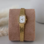 The Petite Mesh Timepiece features a gold diamond bezel and a rectangular, diamond-studded face, elegantly displayed on a beige suede cushion.