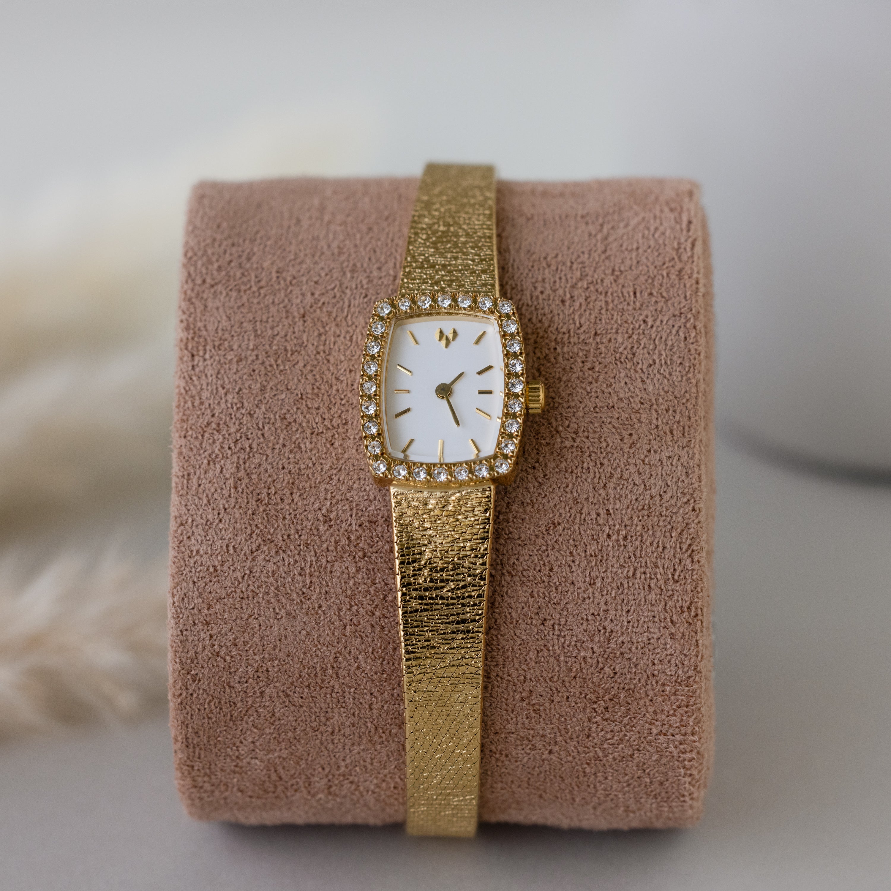 The Petite Mesh Timepiece features a gold diamond bezel and a rectangular, diamond-studded face, elegantly displayed on a beige suede cushion.