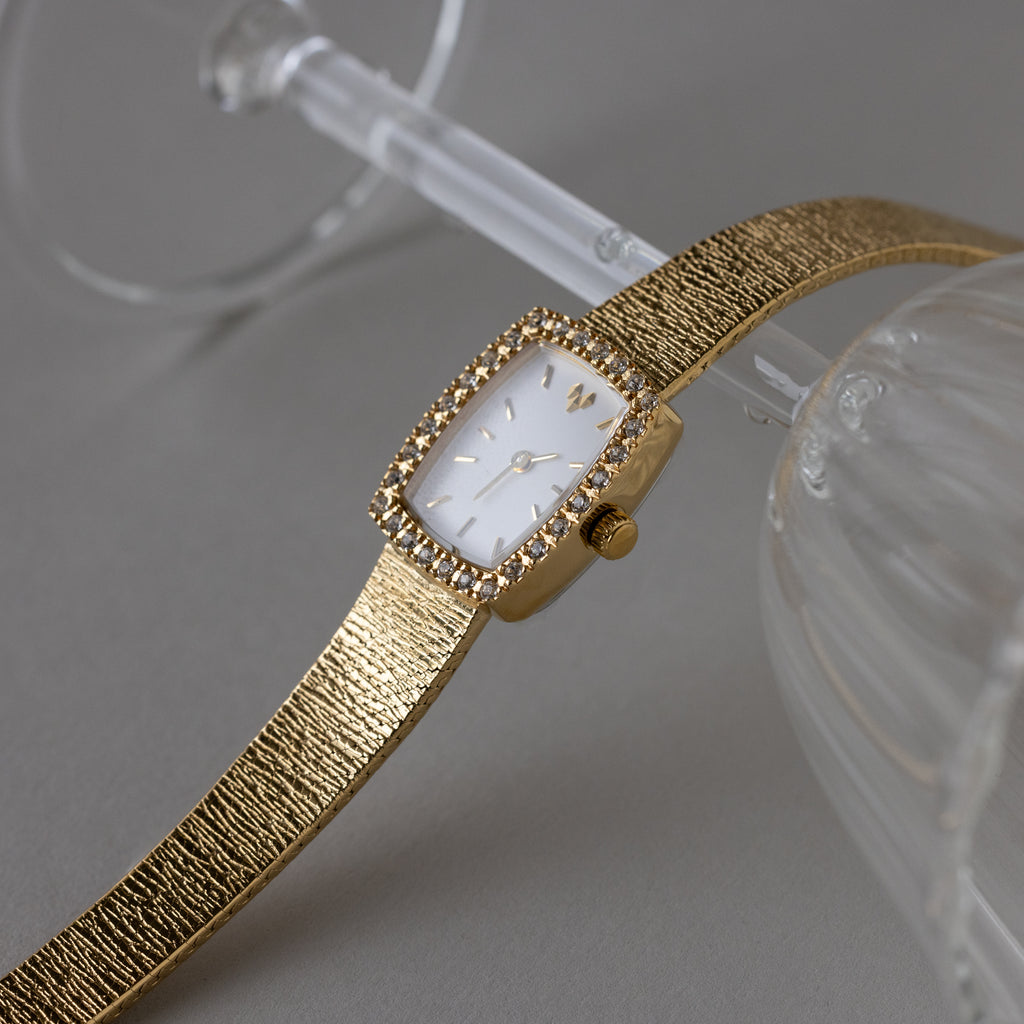 The Petite Mesh Timepiece, featuring a gold rectangular face and textured mesh band, is elegantly displayed on the rim of a glass.