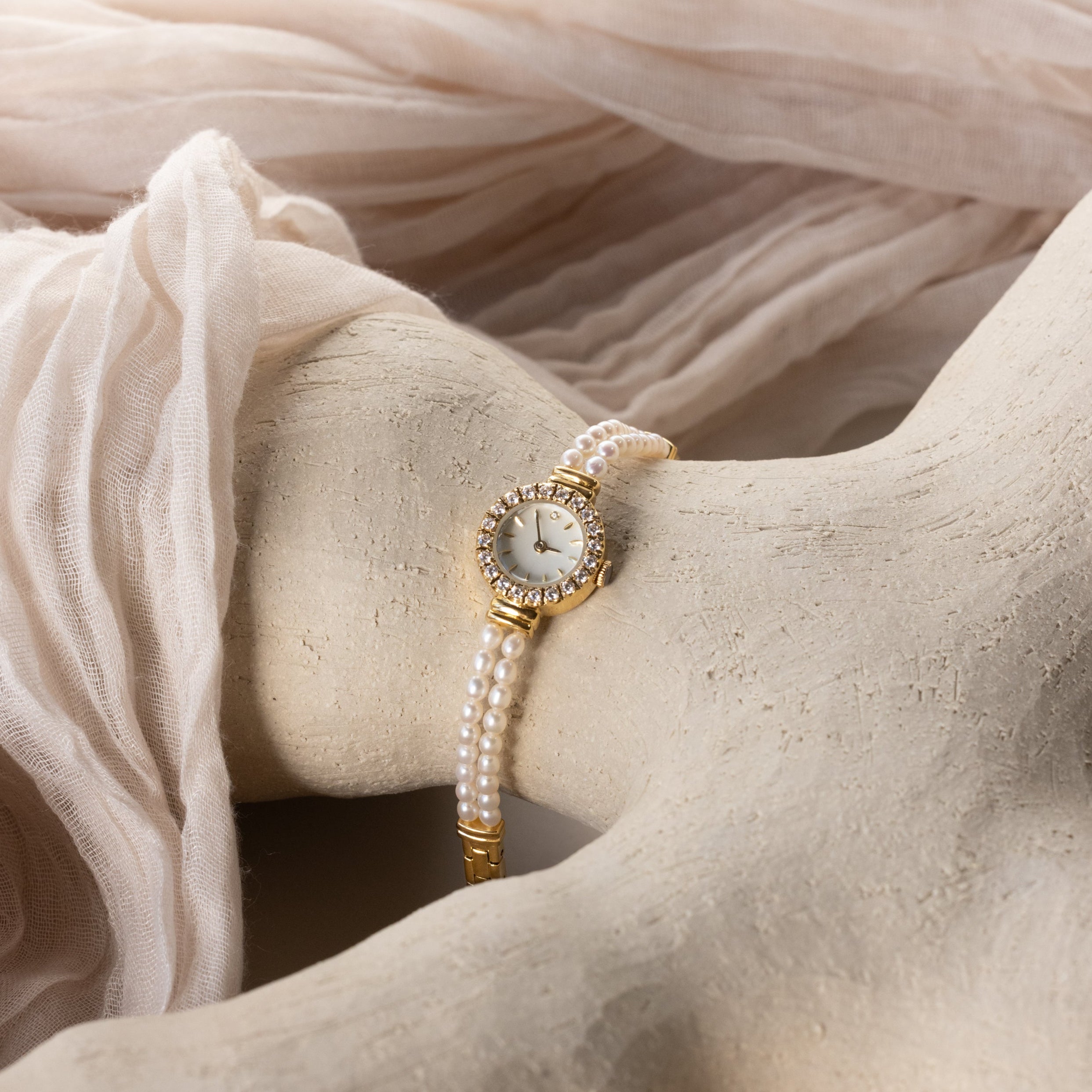 The Pearl Link Timepiece showcases vintage elegance with a jeweled bezel and diamond accents, elegantly displayed on a sculpted white surface with soft beige fabric highlights.