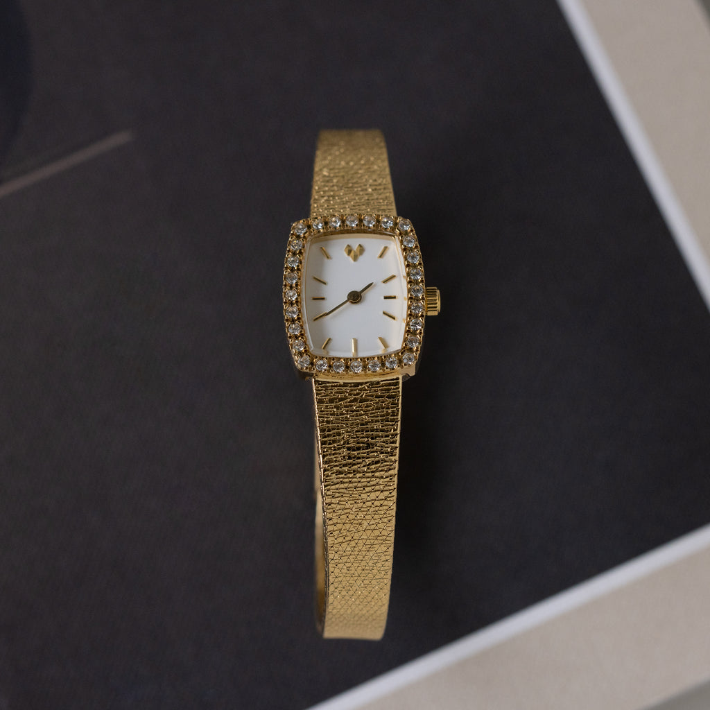 The Petite Mesh Timepiece features a gold rectangular face with diamond-like stones around the dial, shown elegantly on a dark surface.